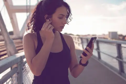 Frau auf einer Brücke, die sich Kopfhörer einsetzt und auf ihr Handy schaut. Sie ist bereit für ihre täglichen 10.000 Schritte.