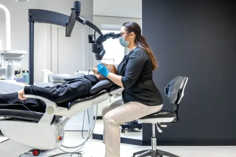 Dentystka w masce i rękawiczkach precyzyjnie pracuje pod mikroskopem, kiedy leczenie kanałowe jest wykonywane z najwyższą starannością.