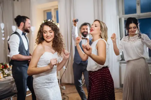 Brautpaar und Gäste tanzen ausgelassen. Frauen im lässigen Chic auf einer Hochzeit, die Braut trägt ein Spitzenkleid.