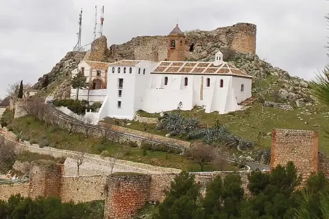 Iglesia Nuestra Señora de Gracia, un edificio blanco con tejado a dos aguas, se alza en una colina rocosa junto a ruinas de un castillo.
