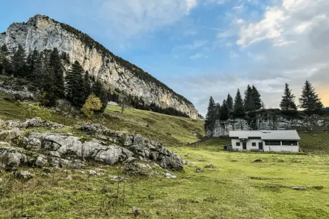 Un chalet blanc se dresse dans un pré verdoyant au pied d'une falaise imposante. Le col de l'Alpette offre un paysage alpin paisible.