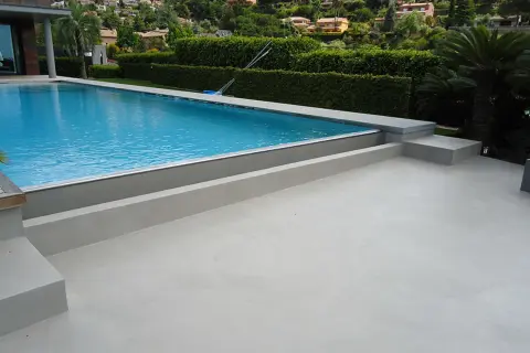 Modern villa with a bright blue pool and surrounding terraces finished with elegant piscinas de microcemento. Lush green hedges and palm trees frame the scene.