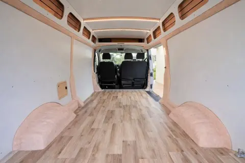 Interior de furgoneta camper con suelo de madera. Las paredes están revestidas y hay asientos en la parte delantera.