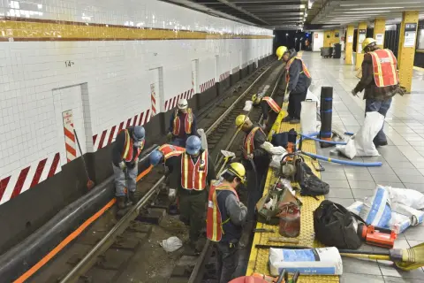 Des ouvriers en tenue de chantier, gilets de sécurité et casques, travaillent sur les voies d'une station de métro. Ils semblent engagés dans des travaux de maintenance, peut-être le prime habillage ou deshabillage de certains éléments.
