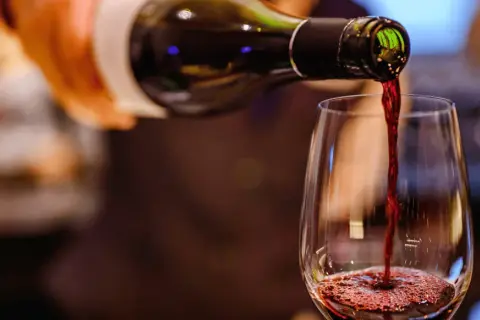 Czerwone wino leje się z butelki do kieliszka, tworząc piankę. Widać bogactwo **tannin w winie**, które nadają mu głębię.