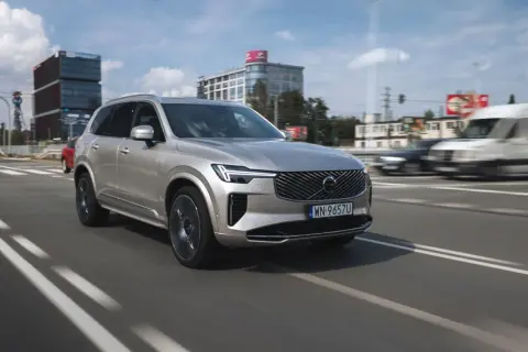 Srebrny Volvo XC90 T8 w ruchu miejskim, z nowoczesnymi budynkami w tle.