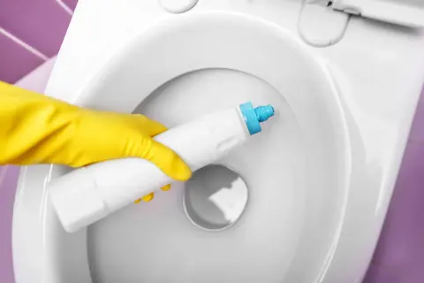 Main jaune appliquant un produit pour éliminer le calcaire dans une toilette blanche.