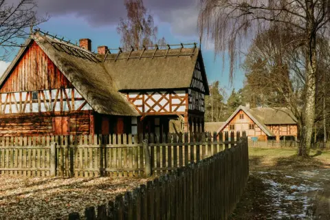 Drewniane domy z krytymi strzechą dachami i drewnianym płotem na tle drzew.