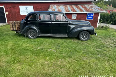 Svart Volvo PV 831 parkerad på gräs framför en röd byggnad med rostigt plåttak. En träkorg sitter på bakre stötfångaren.