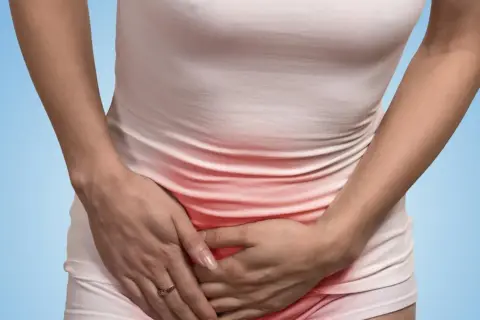 Kobieta trzyma się za brzuch, odczuwając ból w pachwinie. Czerwony obszar na jej ubraniu sugeruje dyskomfort.