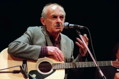 Starszy mężczyzna z gitarą i mikrofonem, śpiewający piosenkę o wojnie.