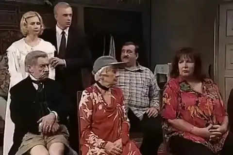 Grupa osób siedzących i stojących w pomieszczeniu, w tym starszy mężczyzna w garniturze i młoda kobieta w białej sukni.