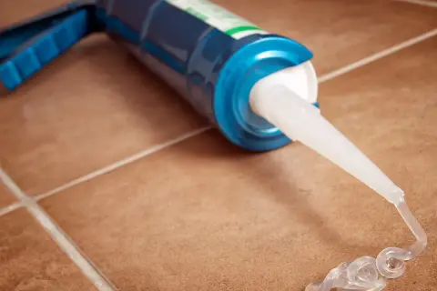 Pistolet do silikonu z wyciśniętym przezroczystym silikonem na brązowych płytkach. Zastanawiasz się, jak długo schnie silikon uniwersalny?