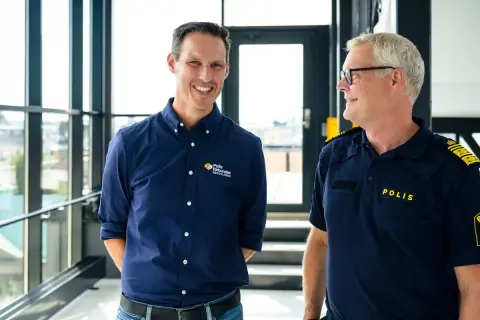 Två män ler mot varandra. Den ena bär en mörkblå skjorta med texten "Polis förbundet Bergslagen", den andra en polisuniform med texten "POLIS".