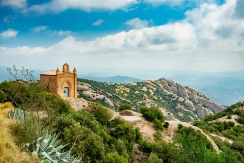 Pequeña ermita en la cima de una montaña, ideal para un **montserrat hike barcelona**. Paisaje rocoso y verde con cielo azul y nubes.