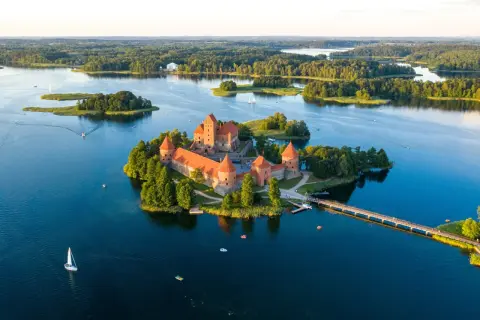 Zamek Troki, malowniczo położony na wyspie pośród jeziora, otoczony zielenią i wodą, z mostem prowadzącym do jego bram.