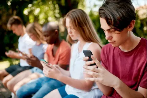 Grupa nastolatków siedzi na ławce, każdy pochłonięty smartfonem. Warto zadbać o ich bezpieczeństwo online, np. poprzez kontrolę rodzicielską na Androidzie.