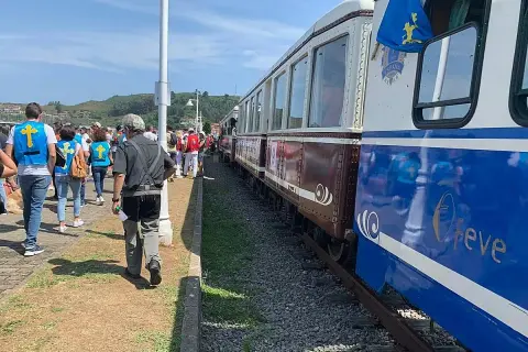 Gente con chalecos amarillos espera el tren descenso del Sella. El tren tiene vagones marrones y azules con detalles dorados.