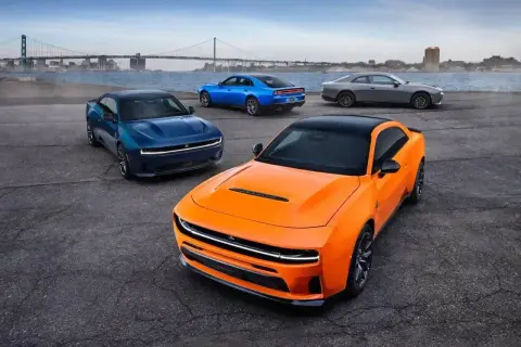 Pomarańczowy i niebieski Dodge Charger Hellcat z 2024 roku, obok szarego coupe, na tle mostu i miasta.