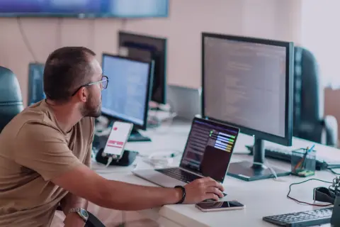Programista aplikacji w okularach pracuje przy biurku z kilkoma monitorami i laptopem.