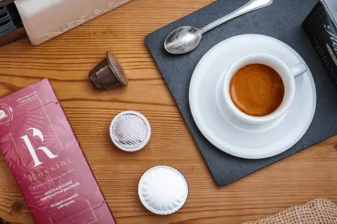 Una tazzina di espresso fumante, capsule e confezione Rossini: un invito a gustare una nota varietà di caffè.