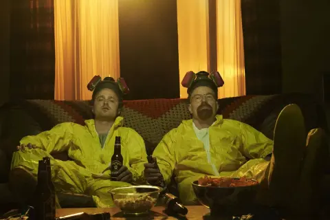 Die Besetzung von Breaking Bad, Walter und Jesse, entspannt in ihren Schutzanzügen mit Bier und Snacks auf dem Sofa.