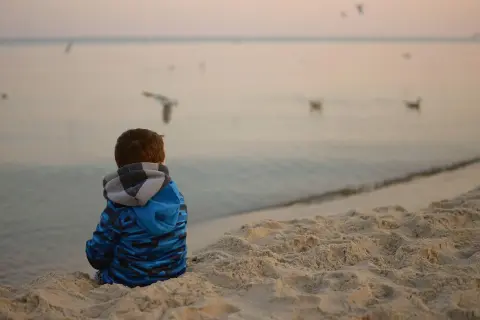 Chłopiec w niebieskiej bluzie siedzi na plaży, obserwując mewy. Spokojna atmosfera nad morzem może pomóc w tym, jak wyciszyć dziecko nadpobudliwe.