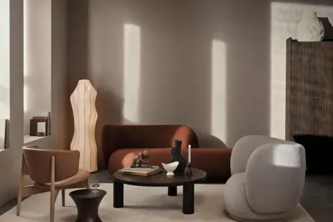 Ein stilvolles Wohnzimmer mit einer rostbraunen Couch. Ein beiger Bouclé-Sessel und ein Holzstuhl ergänzen die Sitzgelegenheiten. Ein runder Couchtisch mit Deko-Objekten steht im Zentrum.