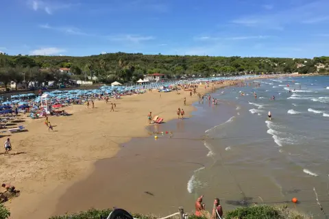 Una giornata di sole sulla **spiaggia di Bescile**, con ombrelloni colorati, bagnanti che giocano e si rilassano, e il mare che invita a un tuffo.