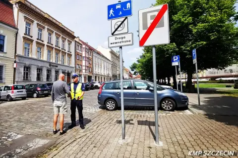Policjant rozmawia z mężczyzną na brukowanej ulicy. Znaki drogowe wskazują strefę zamieszkania i koniec strefy parkowania.