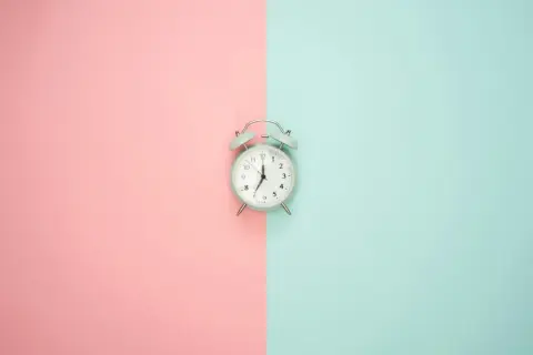 Jasny budzik na tle podzielonym na różowy i turkusowy. Wskazuje godzinę 7:00, więc to poranna godzina am.
