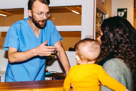 Läkare i blå scrubs pratar med en kvinna som håller ett barn. Han verkar behöva avbryta semester för att hjälpa dem.