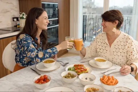 Deux femmes trinquant avec des jus verts et orangés, entourées d'un repas sain. Un exemple de comment ne pas grossir à la ménopause.