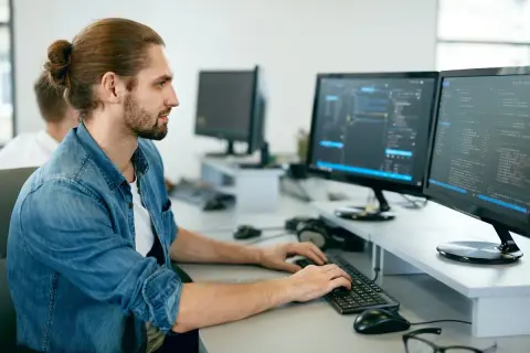 Młodszy programista z kitką na głowie pracuje przy dwóch monitorach wyświetlających kod.