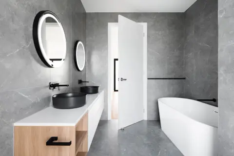 Salle de bain moderne aux murs en **calcaire** gris, avec double vasque noire, baignoire îlot et miroirs lumineux.