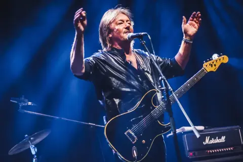 Chris Norman, lider zespołu Smokie, śpiewa i gra na gitarze na scenie, oświetlony niebieskim światłem.
