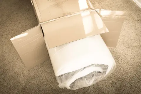 A rolled-up mattress, still in its plastic wrap, sits in an open cardboard box on a carpet. It looks like this left mattress in box too long.