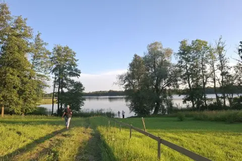Piękny litewski krajobraz nad jeziorem. Osoba idzie ścieżką przez zieloną łąkę w stronę wody, gdzie stoją inni ludzie.