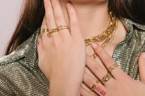 Manos adornadas con varios anillos dorados y un collar grueso, mostrando cómo llevar anillos de manera elegante.