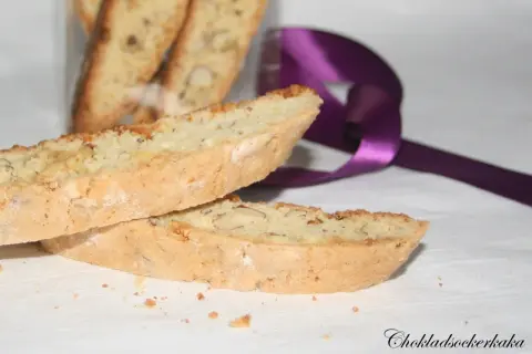 Två mandelkakor, perfekta för ditt biscotti recept, ligger på en vit duk. En lila sidenband ligger bakom.
