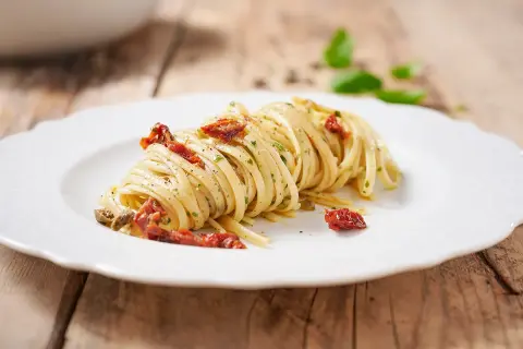 Un piatto di linguine condite con pomodori secchi, capperi e prezzemolo, su un piatto bianco con sfondo in legno.