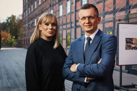 Para adwokatów, kobieta w czarnej bluzce i mężczyzna w niebieskiej marynarce, stoją przed ceglanym budynkiem.