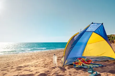 Kolorowy namiot na plaży, obok butelka wody, ręcznik i klapki. Słońce odbija się w morzu.