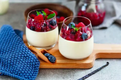 Zwei Gläser cremiger Panna Cotta mit Beerenkompott, ein köstliches rezept panna cotta original italienisch. Blaue Serviette und Holzlöffel mit Blaubeeren.