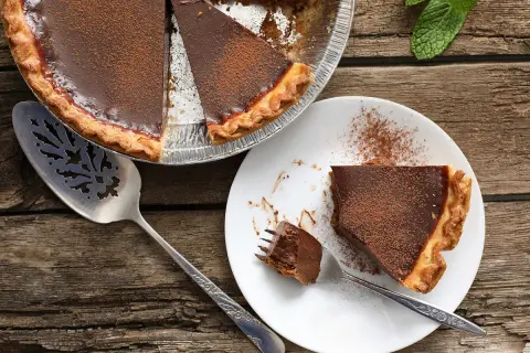 Une délicieuse tarte au chocolat, saupoudrée de cacao, est prête à être dégustée. Une part est servie sur une assiette blanche, avec une fourchette tenant un morceau.