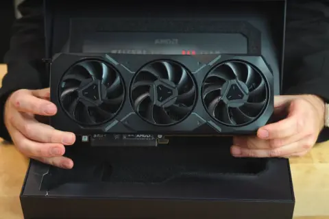 Ręce trzymają kartę graficzną Radeon RX 7900 XTX z trzema wentylatorami, wyjętą z czarnego pudełka z napisem "WELCOME TO RED TEAM".