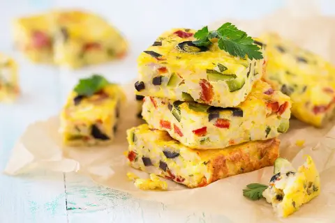 Carrés de frittata colorés, une recette avec des oeufs, garnie de légumes et d'olives, sur du papier sulfurisé.