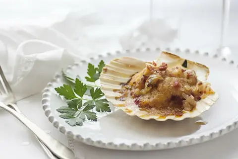 Vieiras a la gallega, gratinadas y servidas en su concha, adornadas con perejil fresco sobre un plato blanco.