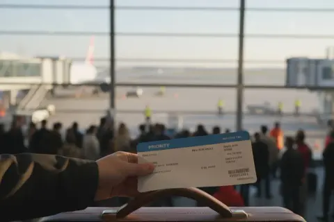 Ręka trzyma kartę pokładową z napisem "PRIORITY" na tle lotniska z samolotem i pasażerami.