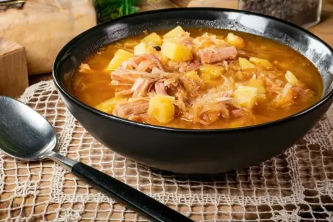 Parzybroda, sycąca zupa z kapusty kiszonej, ziemniaków i mięsa, podana w czarnej misce, obok leży łyżka.